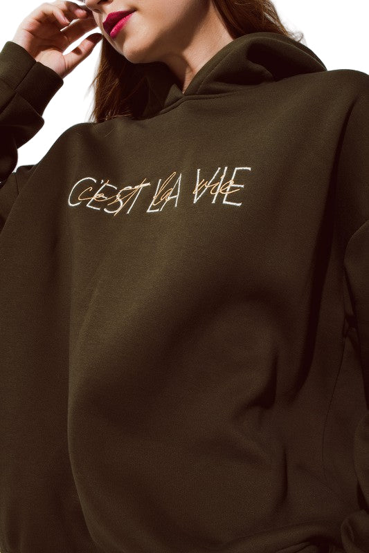Close-up of deep olive “C’est La Vie” hoodie showing soft knit texture and embroidered text on chest.