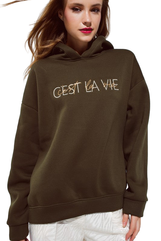 Front view of model in olive colored embroidered “C’est La Vie” hoodie with oversized fit and relaxed sleeves.